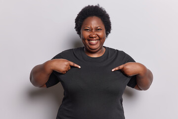 Waist up shot of dark skinned overweightt African woman wears casual black t shirt points at copy space suggests to write your logo here poses against white background. Clothing template concept