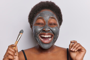 Women and beauty concept. Positive short haired woman with curly hair applies clay facial mask and holds cosmetic brush for applying enjoys skin care procedures exclaims loudly isolated on white wall