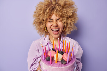 Photo of curly haired young woman poses with delicious cake decorated with macaroons exclaims loudly enjoys celebration wears earrings and shirt isolated over purple background. Holiday concept