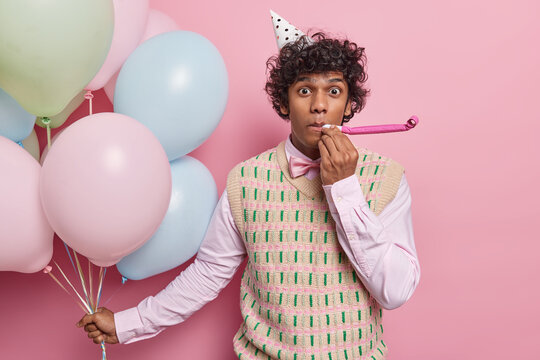 Handsome curly haired Hindu man blows into party horn wears cone hat formal shirt and vest holds bunch of inflated balloons isolated over pink background. People holiday and celebration concept