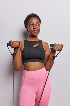 Indoor image of thoughtful dark skinned active woman training in sportswear with fitness equipment working out with resistance band orr expander concentrated aside isolated over white background