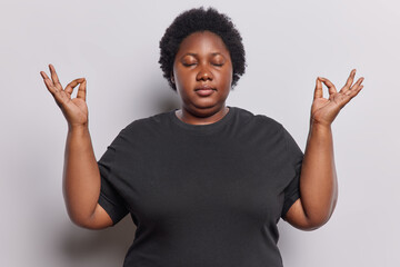 Patience and relaxation concept. Calm dark skinned curly woman keeps eyes closed meditating gathers positive emotions through yoga exercise wears black t shirt isolated over white background.