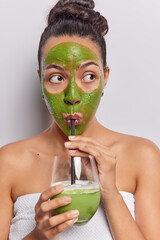Vertical image of Latin woman applies green face mask while savoring smoothie radiating sense of wellnes focused aside has widely opened eyes wrapped in bath towel isolated over white background - obrazy, fototapety, plakaty