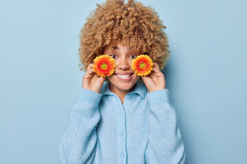 Positive lovely curly haired woman holds orange gerbera flowers near face smiles gladfully wears...