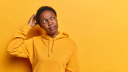 Obraz premium Indoor photo of young thoughtful African american female standing on left isolated on yellow background with blank space for your advertisement keeping one hand on head wearing hoodie looking aside