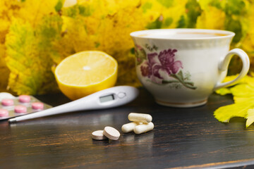 Obraz na płótnie Canvas Capsules, pills, medicine in a blister pack, thermometer, tea cup with lemon on a wooden table with yellow leaves. Concept of treatment autumn cold, flu and coronavirus