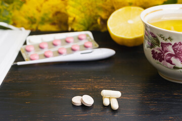 Capsules, pills, medicine in a blister pack, thermometer, tea cup with lemon, on a wooden table with yellow leaves. Concept of treatment autumn cold, flu and coronavirus