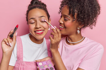 Two stylish friends with curly locks and radiant smiles create captivating makeup magic stand closely enhancing their beauty on pink backdrop prepare for disco party isolated over pink background - obrazy, fototapety, plakaty