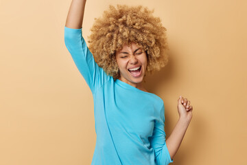 Waist up shot of emotional curly haired woman shakes arms celebrates triumph feels like winner dressed in casual blue jumper keeps eyes closed isolated on brown background. People and success concept