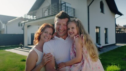 Friendly american dream family demonstrate positive mood, emotions of love and gladness. Parents and daughter are smiling and hugging outdoors near their house. People pose and look at the camera. The