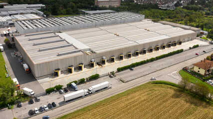Aerial view of a warehouse and its roof. In the parking lot some trucks and vans load and unload the products. Around there are fields and cultivation. - obrazy, fototapety, plakaty