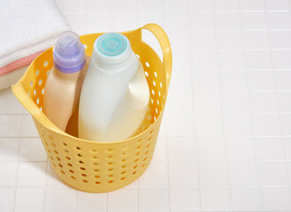 Liquid laundry detergents in bottles stand in a plastic basket. There are clean towels in the background.