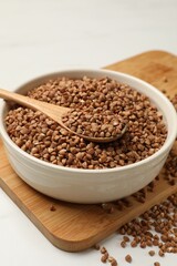 Bowl and spoon with dry buckwheat on white table, closeup