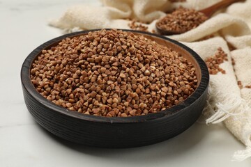 Bowl with dry buckwheat and fabric on white marble table, closeup