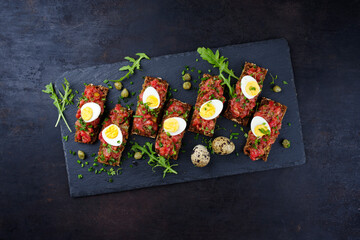 Vollkornbrot Schnitte mit Tartar Belag serviert mit Wachteleiern Kapern und Kräuter als Draufsicht auf einem schwarzen Design Tablett 