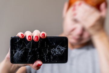 A young woman reacts emotionally to a broken smartphone screen. A pretty woman holds a broken...