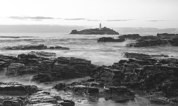 Black and white image of a rocky shoreline with a lighthouse in the background.