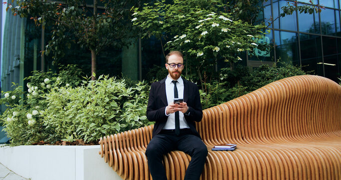 Handsome bearded man using typing mobile phone sitting on bench on city street. Attractive businessman on the phone in an office building. Businessman texing on phone.