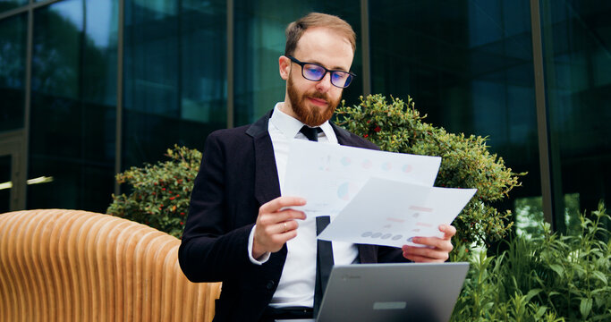 Businessman Reading Documents While Sitting On Bench Outdoor. Man Working On Laptop Remotely From Office Wearing Suit Cheking Document Doing Job. Freelance Worker, Project On The Go Outdoor.