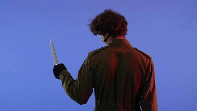 Man with big sharp knife on blue background with red neon light. Image of Jason Voorhees maniac from Friday the 13th. Back view.
