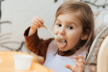 Small cute little toddler brunette caucasian girl with two tails tasting and enjoying by herself with a spoon the greek yogurt sitting in baby chair with messy face self-feeding concept