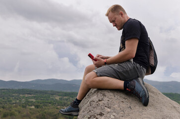 Naklejka na ściany i meble Thoughtful man sitting on a rock on top of mountain peak looking at cell phone screen in hand. Concept of person connected to internet everywhere. Beautiful sky on a cloudy day. Freedom and adventure