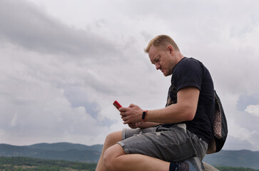 Thoughtful man sitting on a rock on top of mountain peak looking at cell phone screen in hand. Concept of person connected to internet everywhere. Beautiful sky on a cloudy day. Freedom and adventure