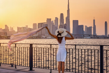 Fototapeta na wymiar A happy tourist woman waving her scarf in the wind looks at the view of the modern skyline of Dubai, UAE, during sunset time
