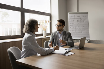 Serious younger businessman talking to mature colleague at laptop, discussing work strategy,...