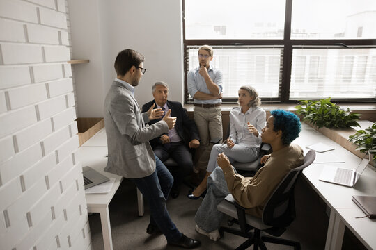 Multiethnic different aged business team brainstorming in office co-working space, sitting in circle, talking, discussing ideas, listening to young male leader, boss