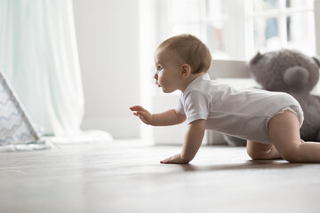 Side view cute pretty girl or baby boy in bodysuit crawling on warm floor in cozy living room. Little toddler exploring surroundings, creeping alone in modern playroom. Development, growth, babyhood