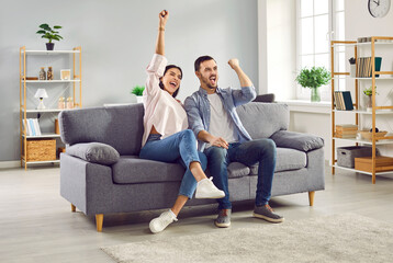 Excited couple watching football or hockey match together at home. Overjoyed happy couple of man and woman raising arms and screaming, celebrating victory of their favorite team - obrazy, fototapety, plakaty