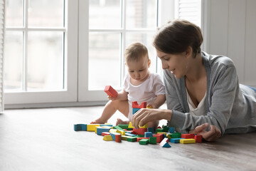 Attractive loving mother play with infant toddler on warm floor, young mum help build tower from...