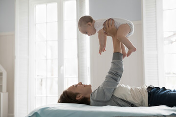 Side view loving mother lying on comfortable bed in cozy, light bedroom play with infant, lifts un...