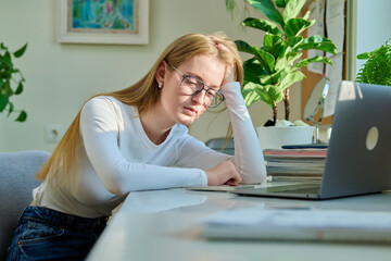 Sad pensive young teenage girl sitting near smartphone laptop at home.