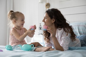 Mother lying on bed in bedroom spend time with cute little daughter, play together, drink morning...