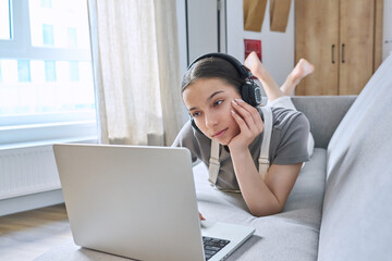 Teen girl in headphones at home on couch using laptop