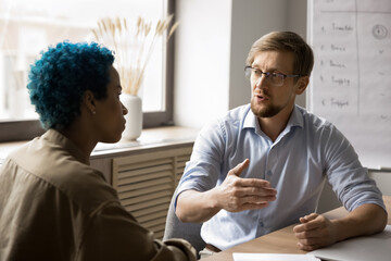 Two multiethnic coworkers talking at office meeting table, discussing work process, collaboration, partnership, planning teamwork. Young business leader man, boss explaining task to colleague woman - obrazy, fototapety, plakaty