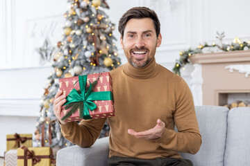 Obraz na płótnie Canvas Man sitting at home celebrating New Year and Christmas holidays, using laptop for video call, showing gift box to webcam, greeting friends and family in living room remotely from home
