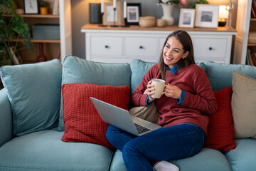 Obraz premium Smiling woman freelancer sitting on sofa in home office with coffee cup and laptop.
