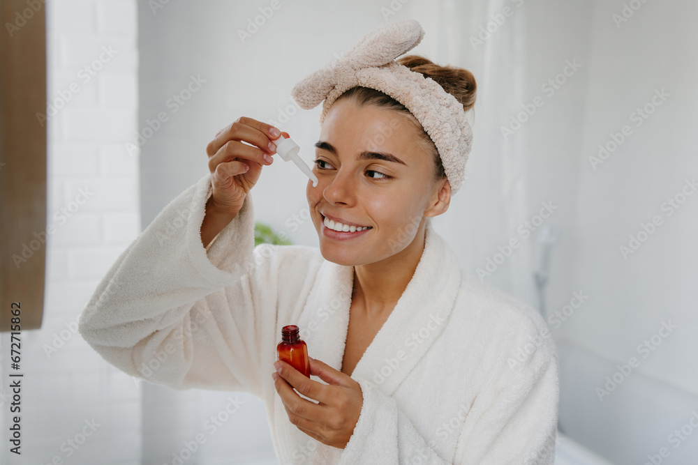 Wall mural happy young woman in bathrobe applying cosmetic serum on face while standing in bathroom