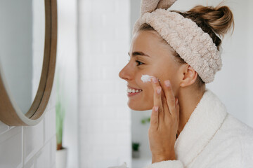 Attractive young woman in bathrobe applying face cream and smiling while standing in bathroom