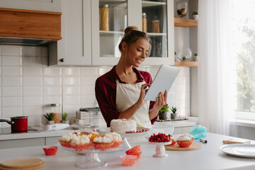 Smiling female pastry chef using digital tablet while preparing cakes and muffins at the domestic kitchen