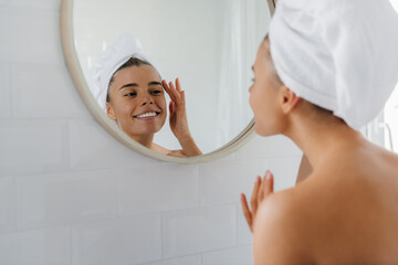 Happy young woman with towel over head touching face while looking at the mirror in bathroom