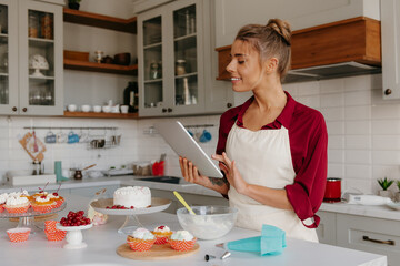 Confident female confectioner using digital tablet while preparing cakes and muffins at the kitchen