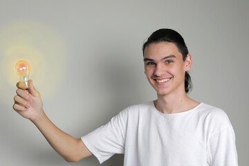 Teenager boy with long hair smiles holding glowing yellow light bulb in his hand