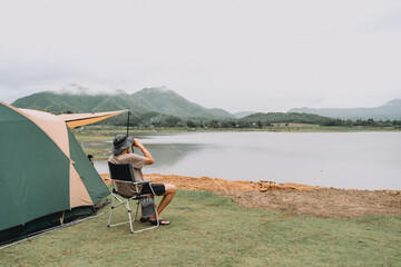 Male tourist sitting next to tent, sitting on camping chair, looking at morning view with lake and mountains.