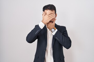 Handsome business hispanic man standing over white background covering eyes and mouth with hands, surprised and shocked. hiding emotion