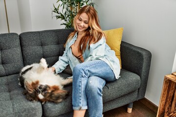 Young caucasian woman sitting on sofa playing with dog at home - obrazy, fototapety, plakaty