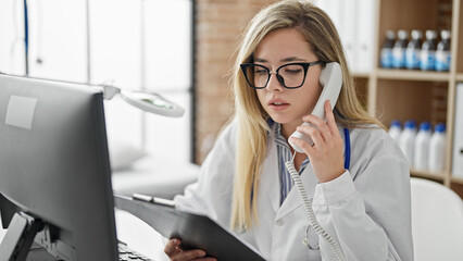 Young blonde woman doctor talking on telephone reading medical report at clinic